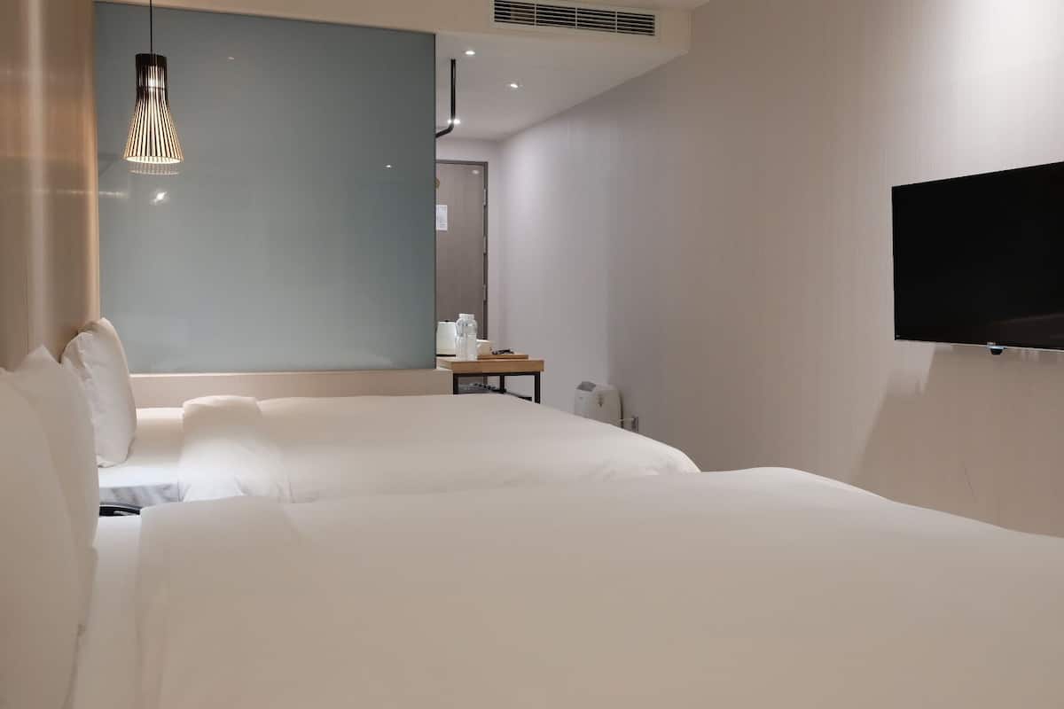A hotel room with two large beds, a flat-screen TV mounted on the wall, a bedside table with a lamp, and a mirror on the wall.