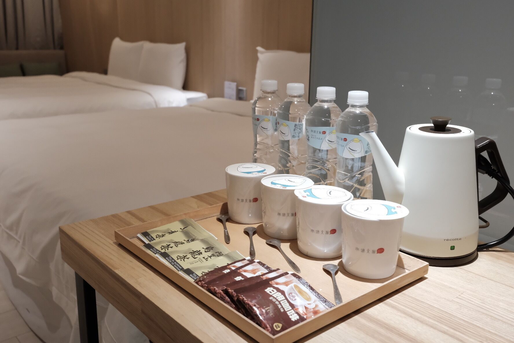 Una habitación de hotel con una cama, una mesita de madera con bolsitas de té, botellas de agua y una tetera.