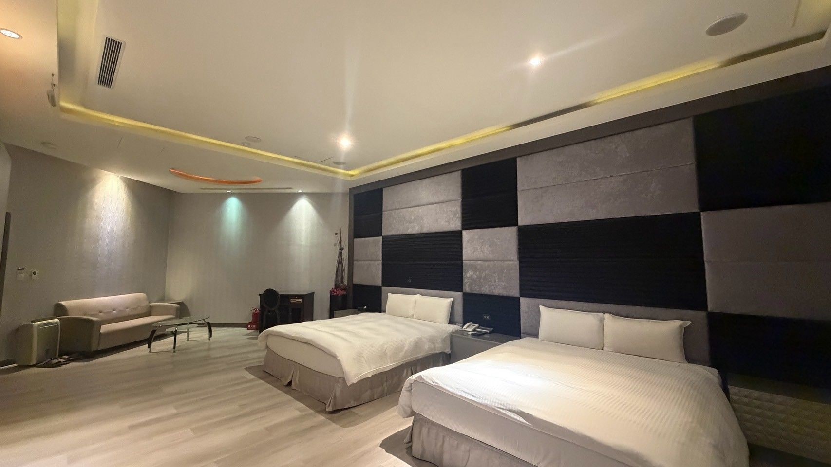 Quarto de hotel moderno com duas camas, um sofá, uma mesa pequena e um painel decorativo na parede.