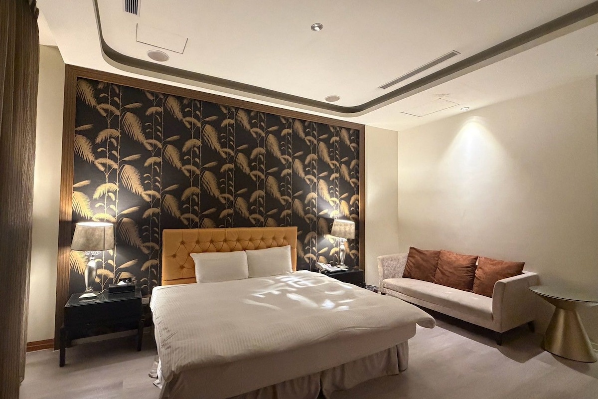 Een hotelkamer met een groot bed, een slaapbank, een nachtkastje met lampen en behang met een patroon.