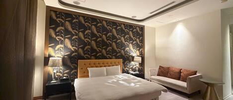 Quarto de hotel com uma cama grande, um sofá, uma mesinha de cabeceira com luminárias e um papel de parede estampado.