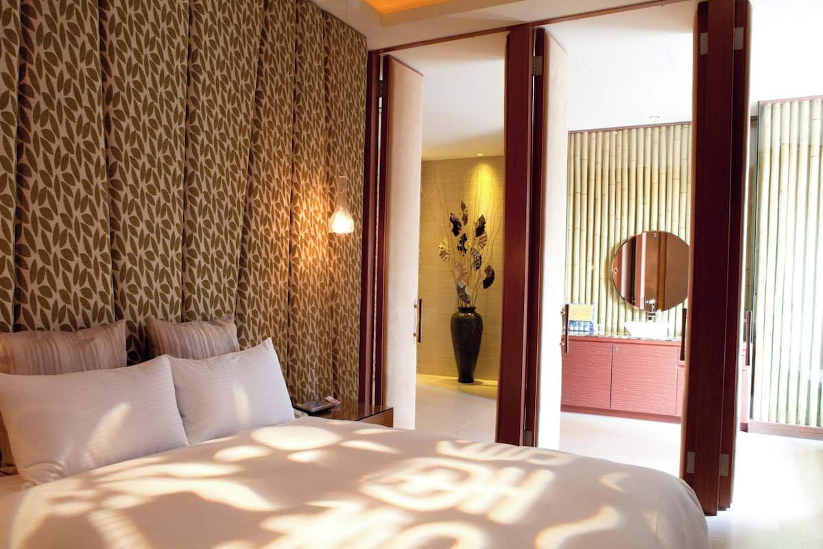 Une chambre avec un mur à motifs, un lit avec des oreillers, une table de chevet et une vue sur une salle de bain avec un miroir et un vase.