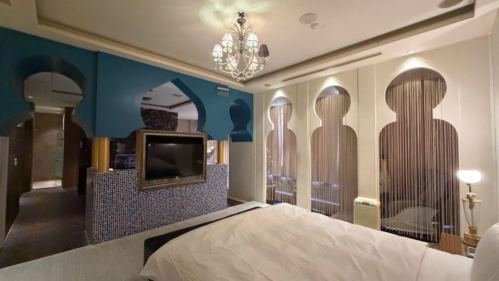 Une chambre d’hôtel avec un grand lit, un téléviseur fixé au mur, un lustre et un miroir à rayures verticales.