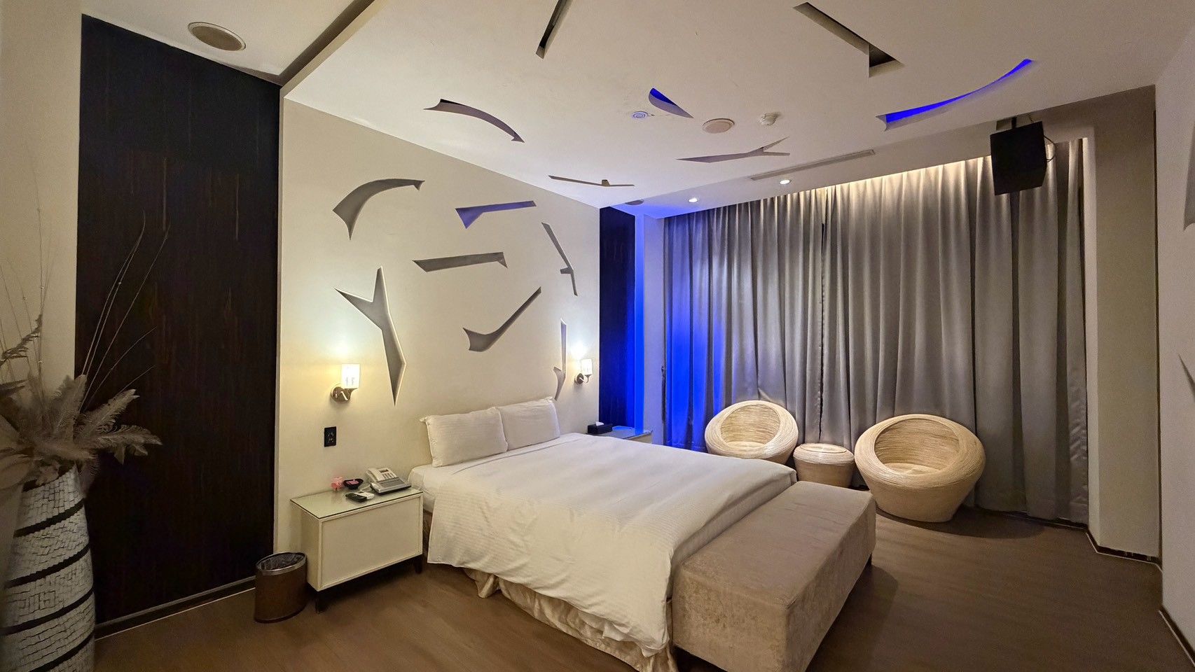 A modern hotel room with a large bed, bedside tables, a wall-mounted TV, and a seating area with two chairs.