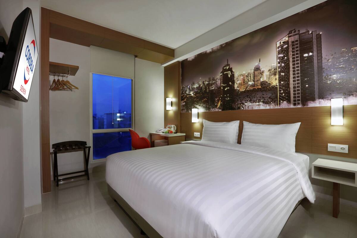 A hotel room with a large bed, a cityscape mural, a television, and a window with a view of the city at night.