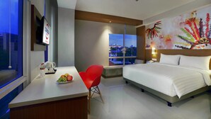 Une chambre d’hôtel moderne avec un grand lit, un bureau doté d’une machine à café, un fauteuil rouge et une vue sur la ville par la fenêtre.