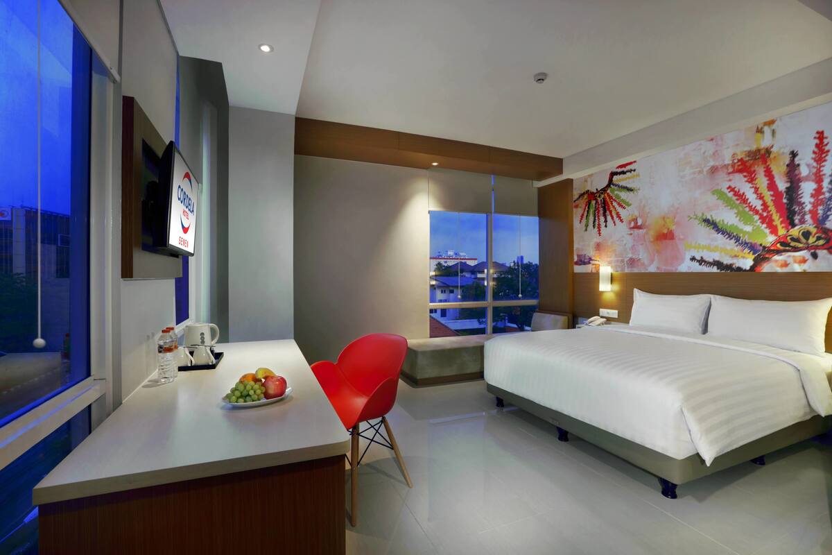 A modern hotel room with a large bed, a desk with a coffee maker, a red chair, and a city view through the window.