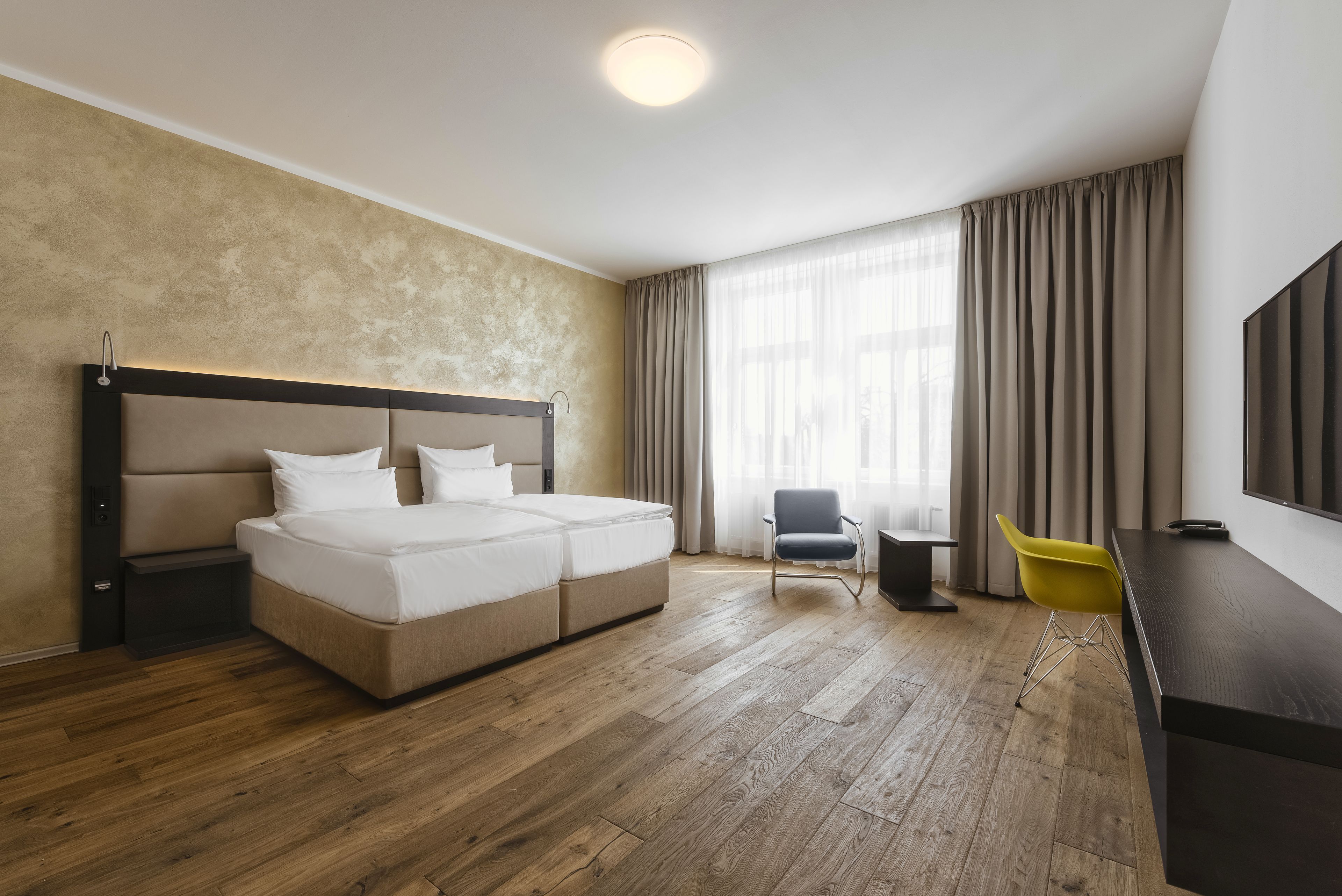 A hotel room with a double bed, a desk with a chair, a TV, and a large window with curtains.