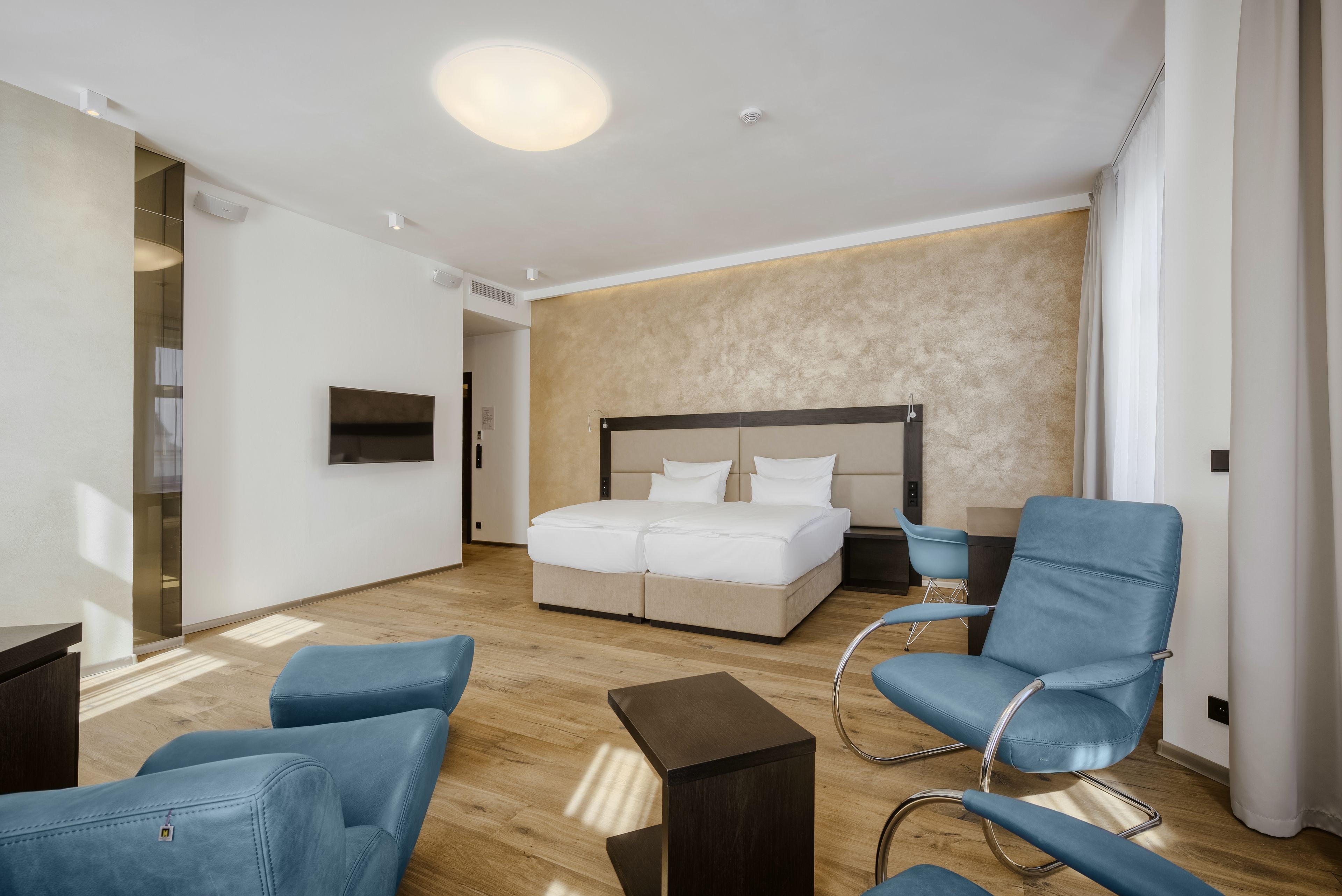 A modern hotel room with a large bed, two blue armchairs, a small table, and a TV mounted on the wall.