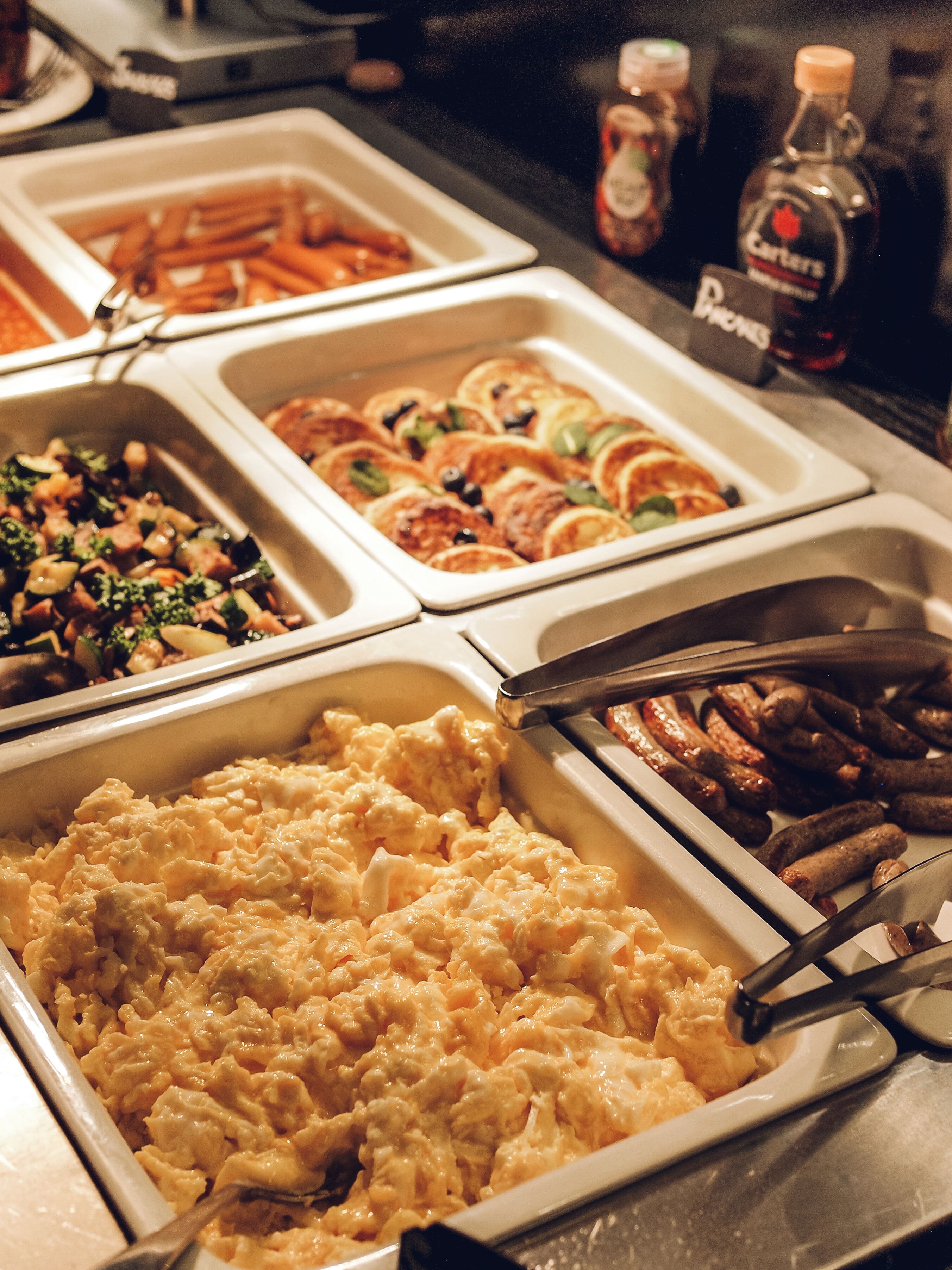 Buffet com diversos pratos, incluindo ovos mexidos, linguiça e um prato gratinado.