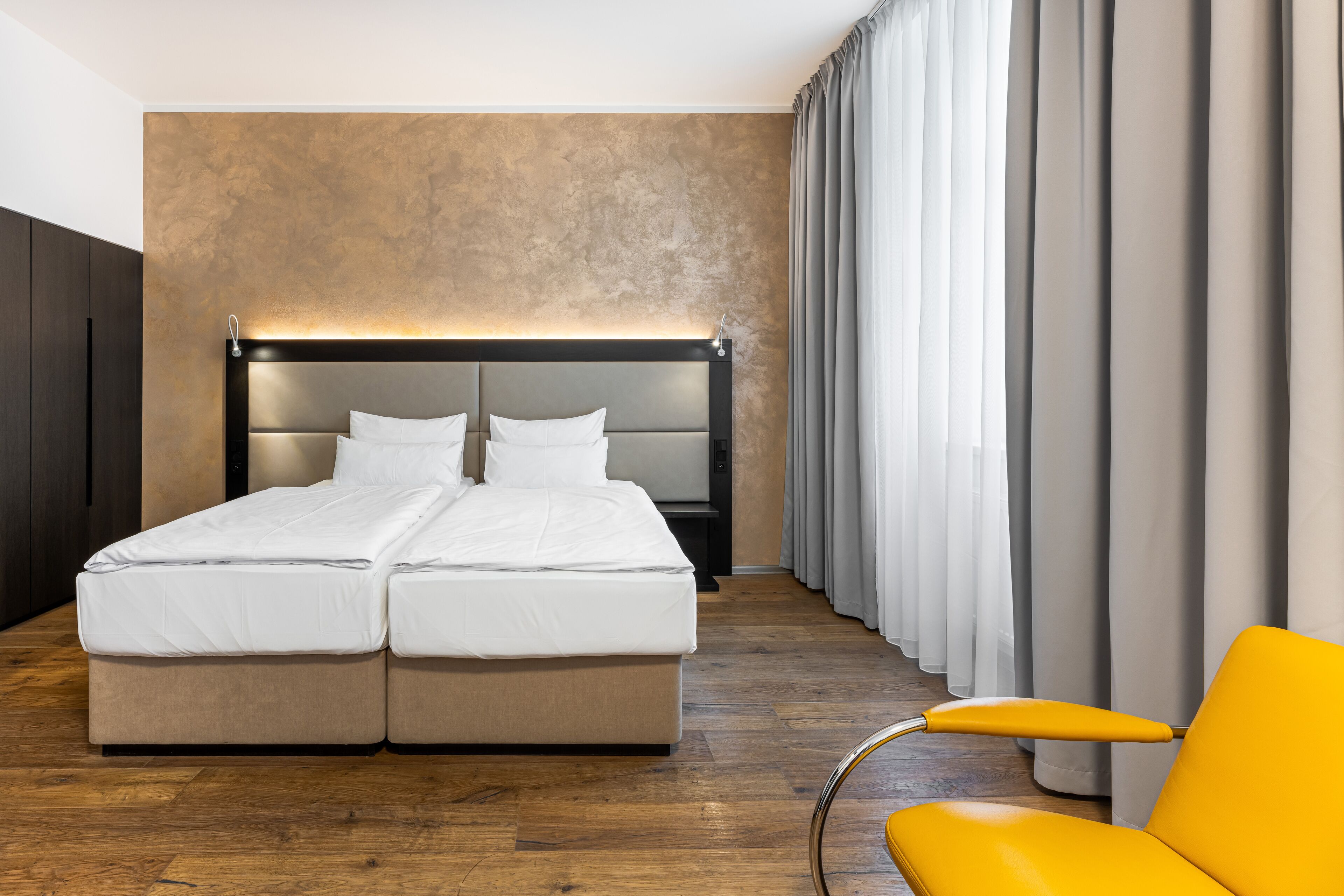 Una cama doble con cabecera, un sillón amarillo y un ventanal con cortinas.