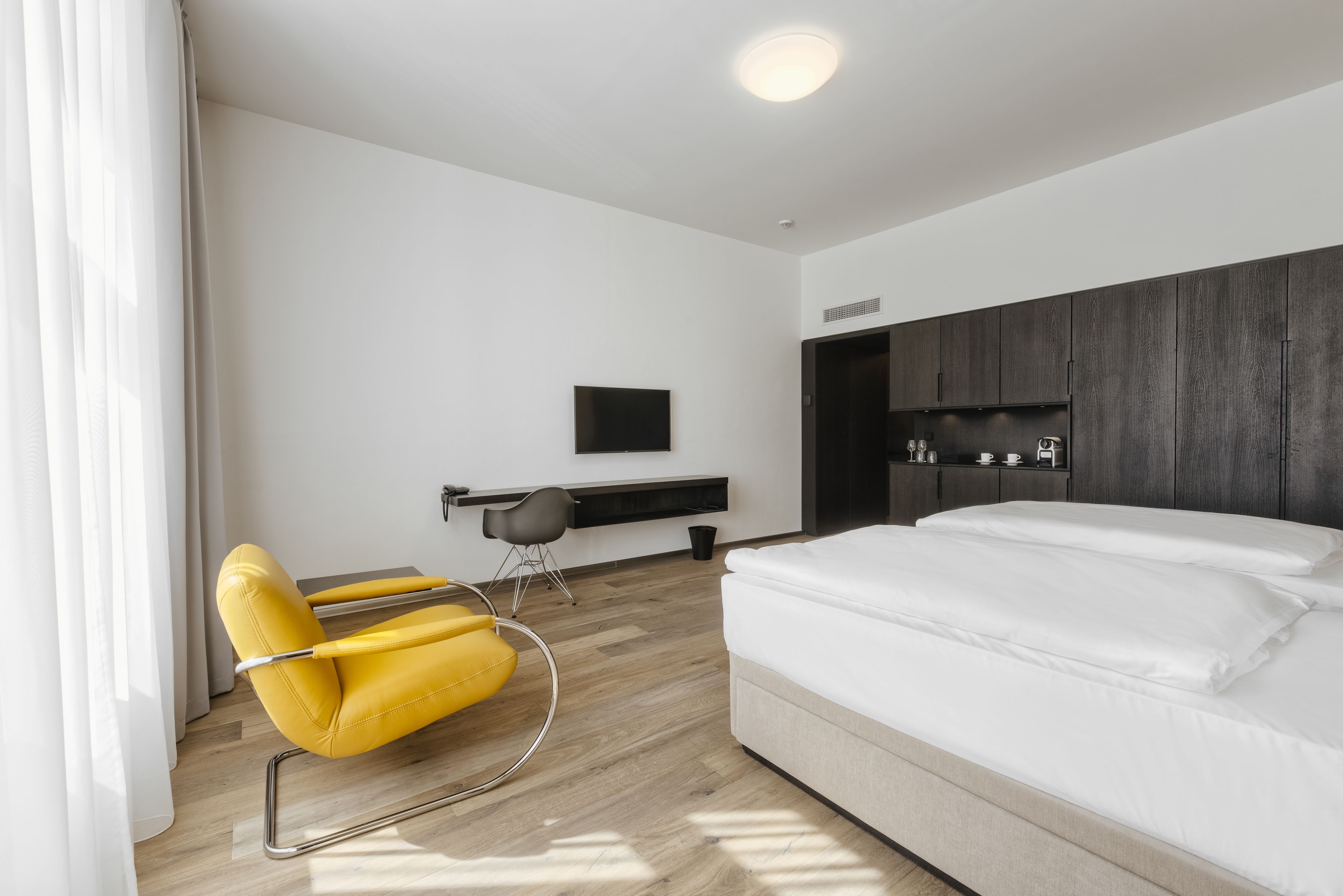 A modern hotel room with a bed, a desk, a chair, and a TV on the wall.