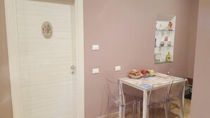 Un comedor con una mesa dispuesta para cuatro, una puerta blanca con un adorno y una repisa montada en la pared con diversos objetos.