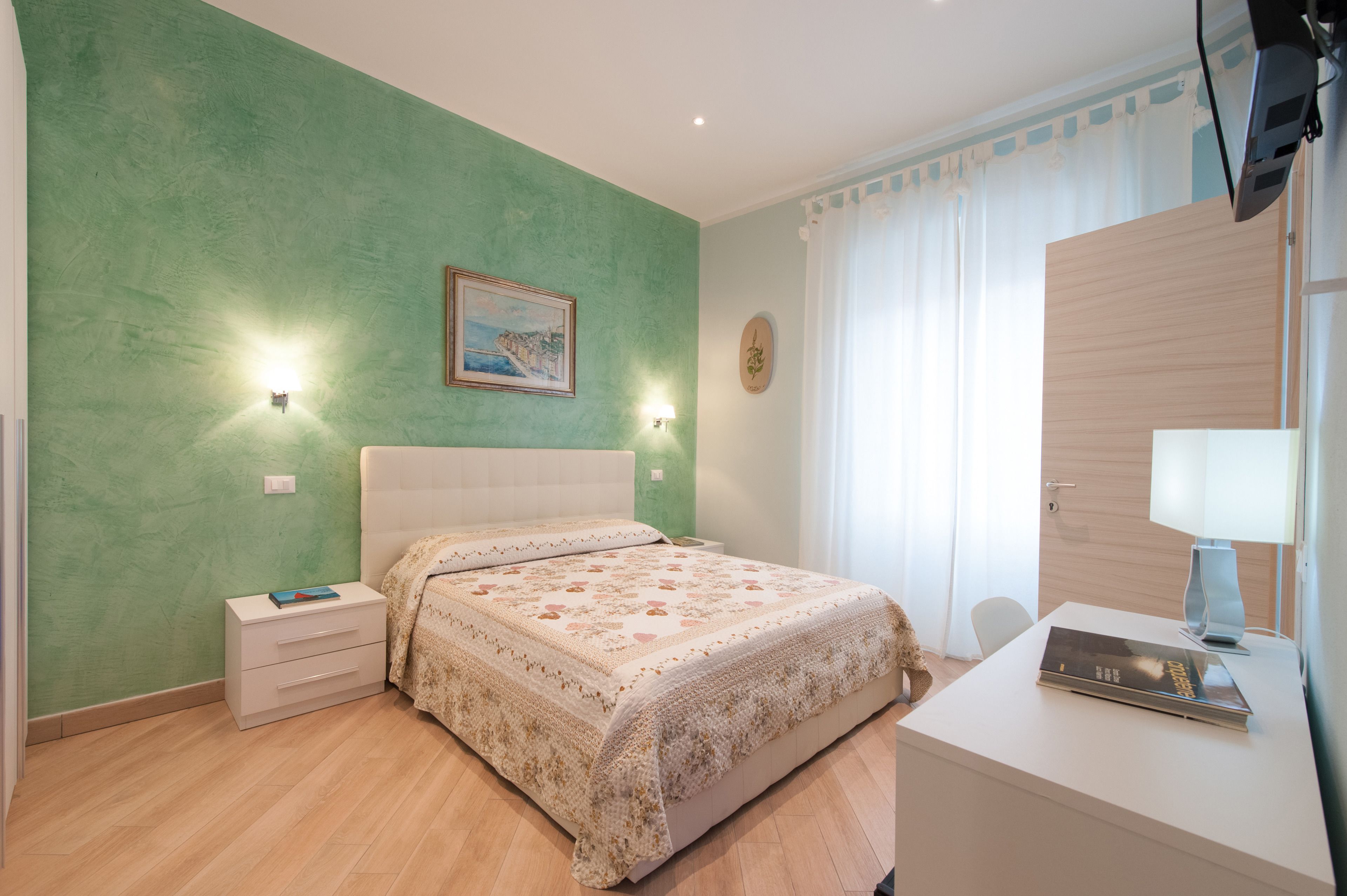 Quarto com cama, mesas de cabeceira, escrivaninha com luminária, televisão e janela com cortinas.