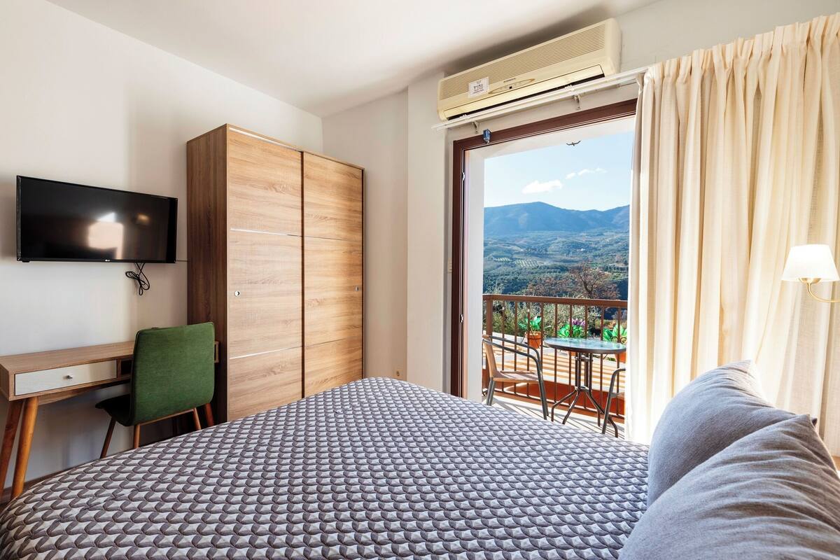 A hotel room with a bed, a desk, a chair, a TV, an air conditioner, and a balcony with a view of the mountains.