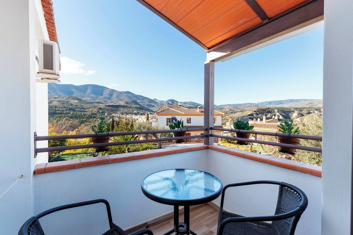 A balcony with a glass table and two chairs, overlooking a mountainous landscape with a view of a building with a tiled roof.