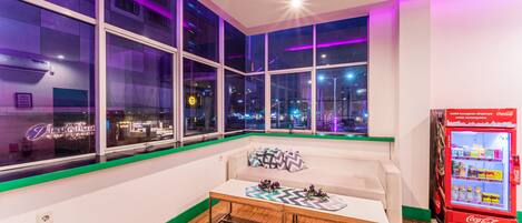 A modern hotel room with large windows, a white sofa, a wooden table, and a Coca-Cola refrigerator.