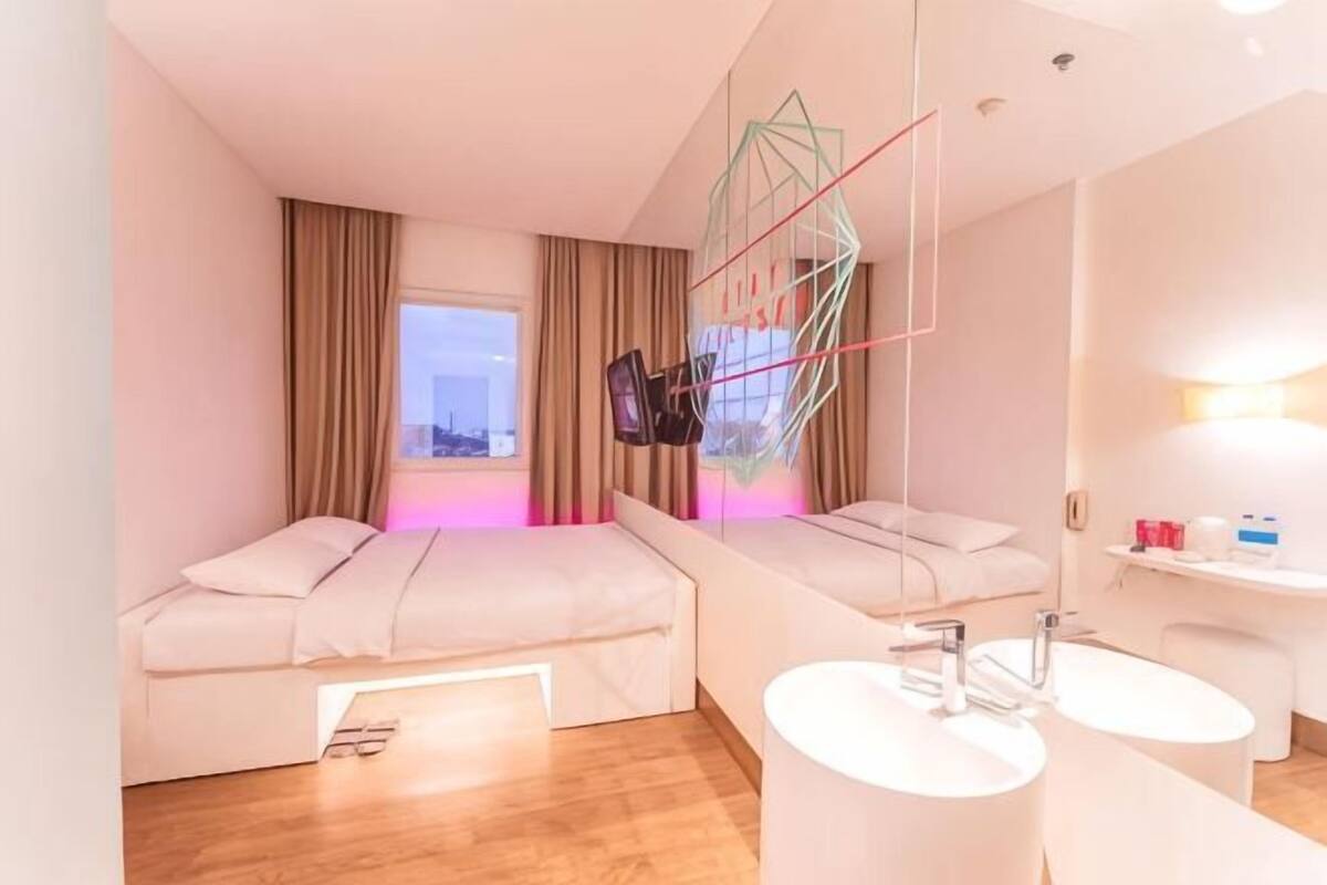 A hotel room with two beds, a large window, a bathroom with a sink and mirror, and a city view.