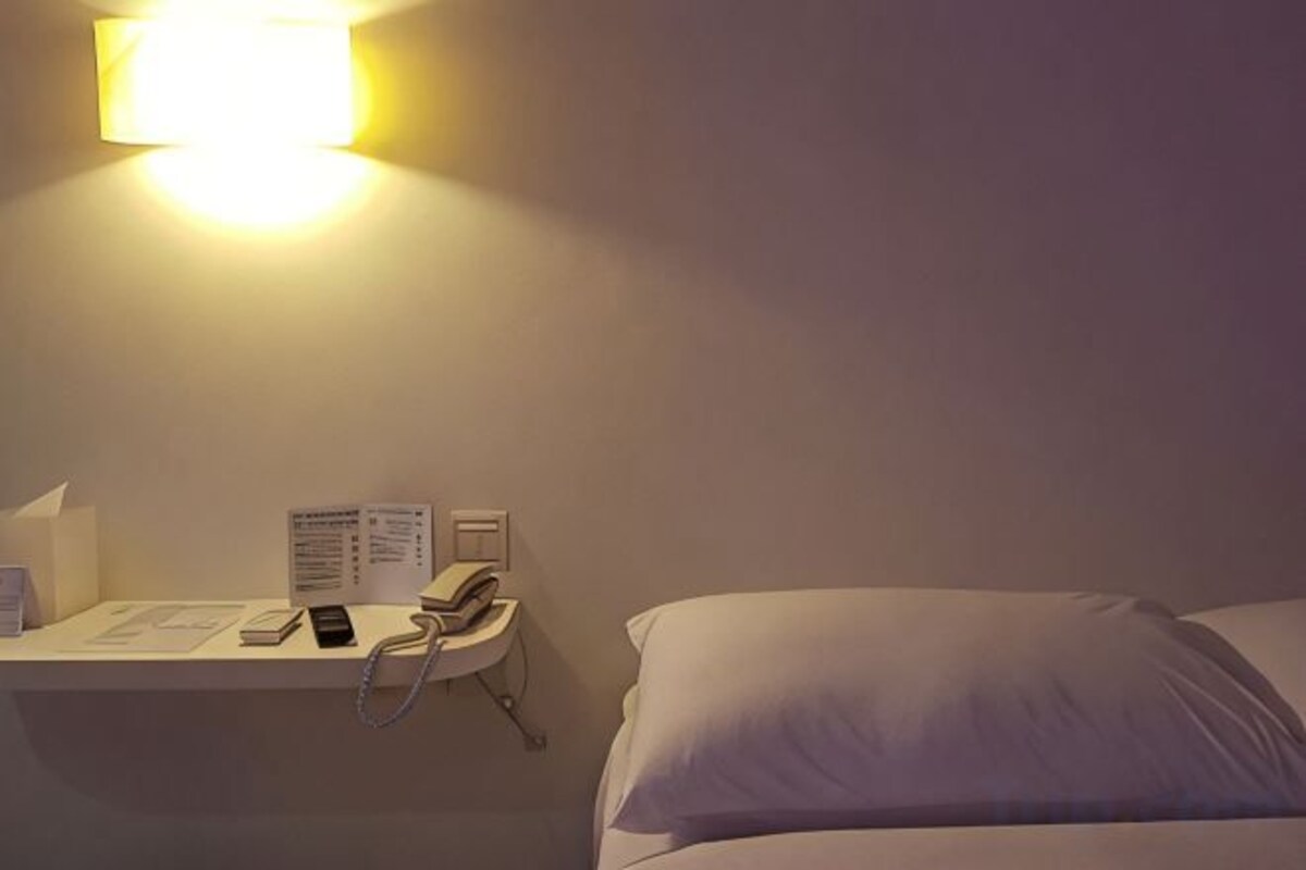A hotel room with a bed, a bedside table with a phone, and a wall-mounted light fixture.