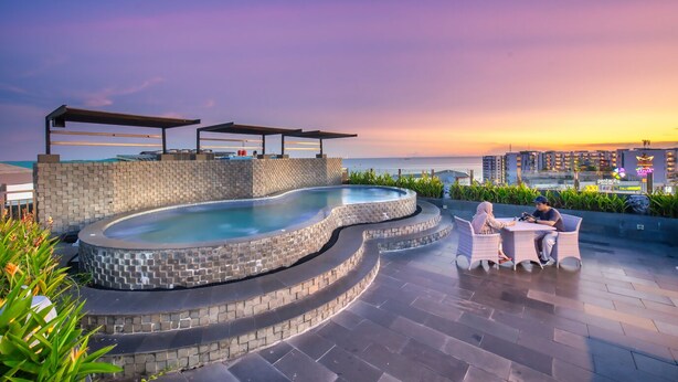 Ein Pool auf dem Dach mit geschwungener Form, umgeben von einer Steinmauer und mit Blick auf eine Stadtlandschaft bei Sonnenuntergang.