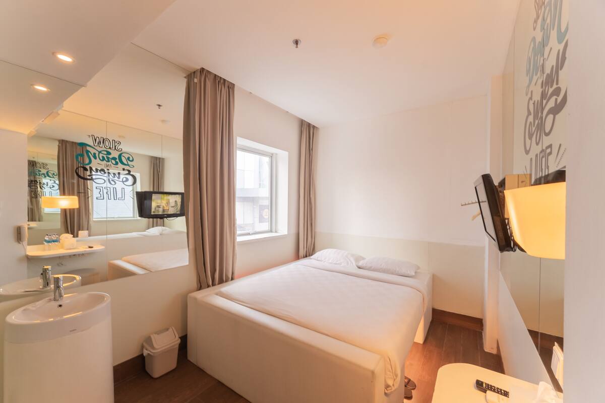 A hotel room with a bed, a bedside table, a TV, a window with curtains, a bathroom with a sink and mirror, and a wall with motivational text.
