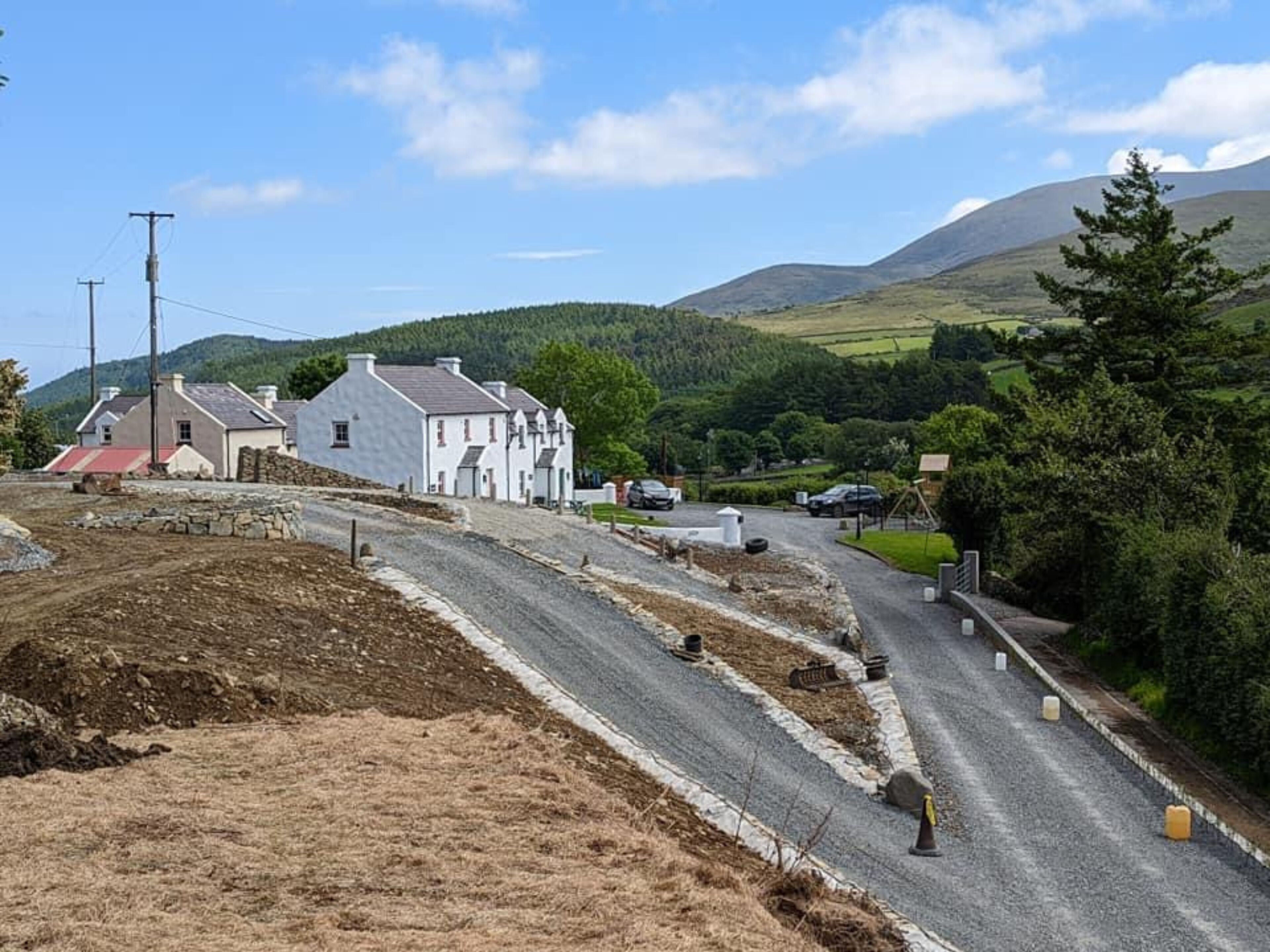 Une route de campagne, bordée de maisons d’un côté, avec un paysage vallonné en arrière-plan.