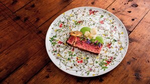 A plate with a slice of meat, green leaves, and small flowers on a wooden table.