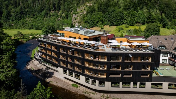 A multi-story hotel with balconies overlooking a river, surrounded by forested hills.