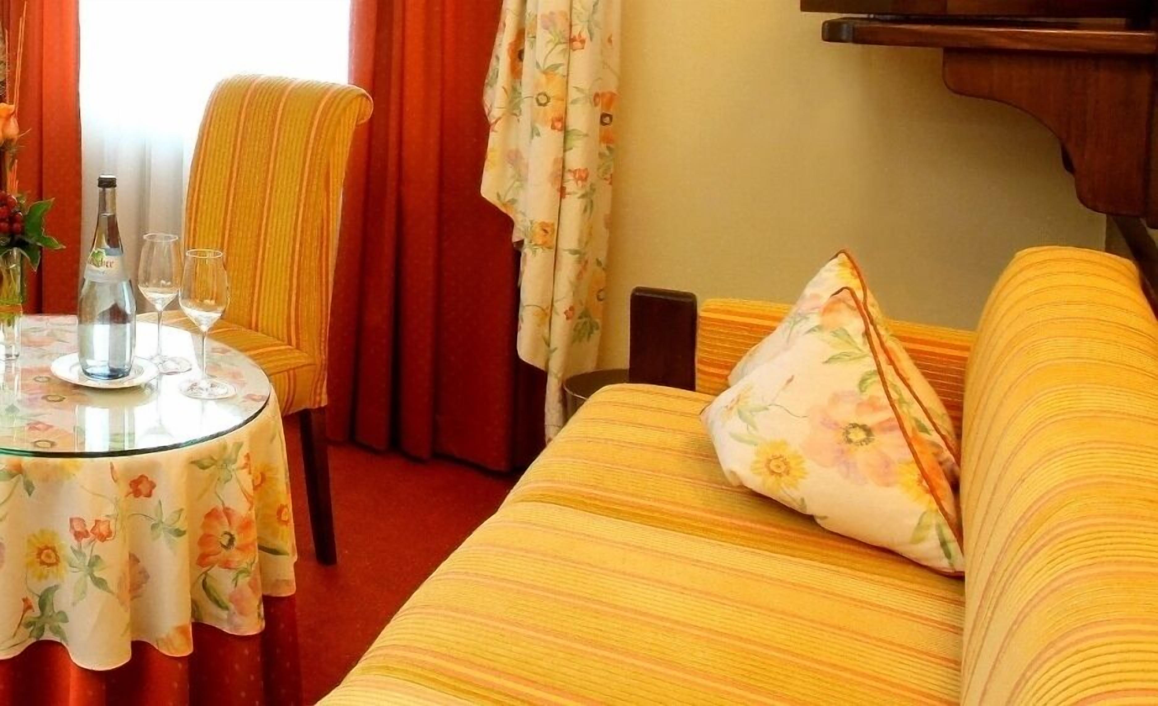 Ein Hotelzimmer mit einem gestreiften gelben Sofa, einem runden Glastisch mit einer blumigen Tischdecke, einem Sessel mit einem gelb-gestreiften Kissen und einem roten Vorhang.