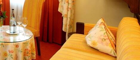 Habitación de hotel con un sofá a rayas amarillas, una mesa redonda de vidrio con mantel floral, una silla con cojín amarillo rayado y una cortina roja.