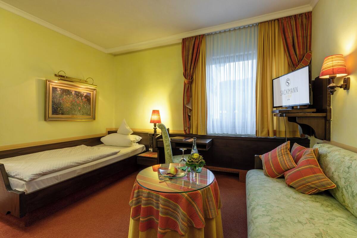 Ein Hotelzimmer mit einem Bett, einem Sofa, einem kleinen Tisch mit Lampe, einem Fernseher und einem Bild an der Wand.