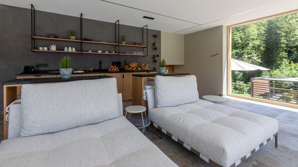 A modern living room with a sofa set, a kitchen area with wooden shelves, and a view of a balcony with greenery. - Sackmann Schwarzwald Charme (Baiersbronn)