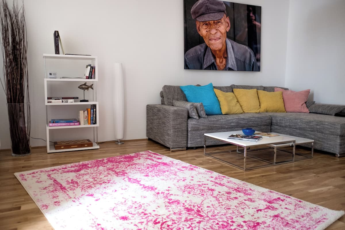 Una sala de estar con un sofá modular gris, cojines de colores, una estantería blanca y una alfombra estampada rosa.