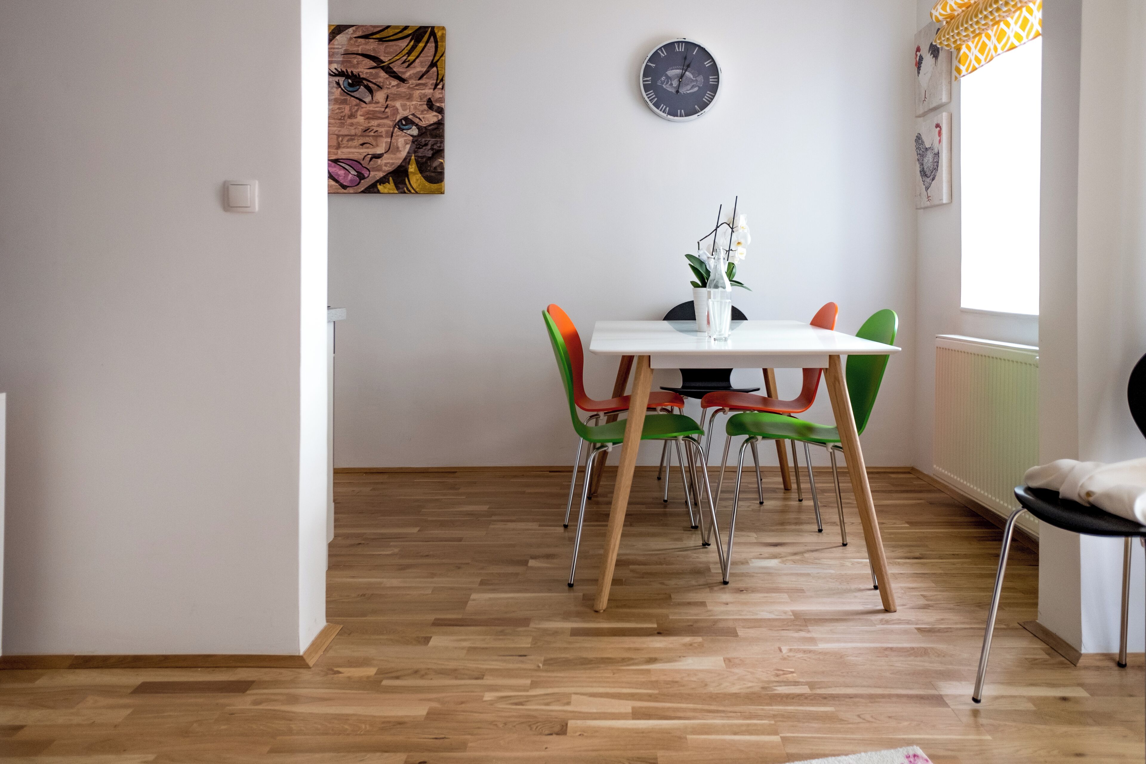 Un comedor con una mesa blanca, sillas coloridas, un reloj y obras de arte en las paredes.