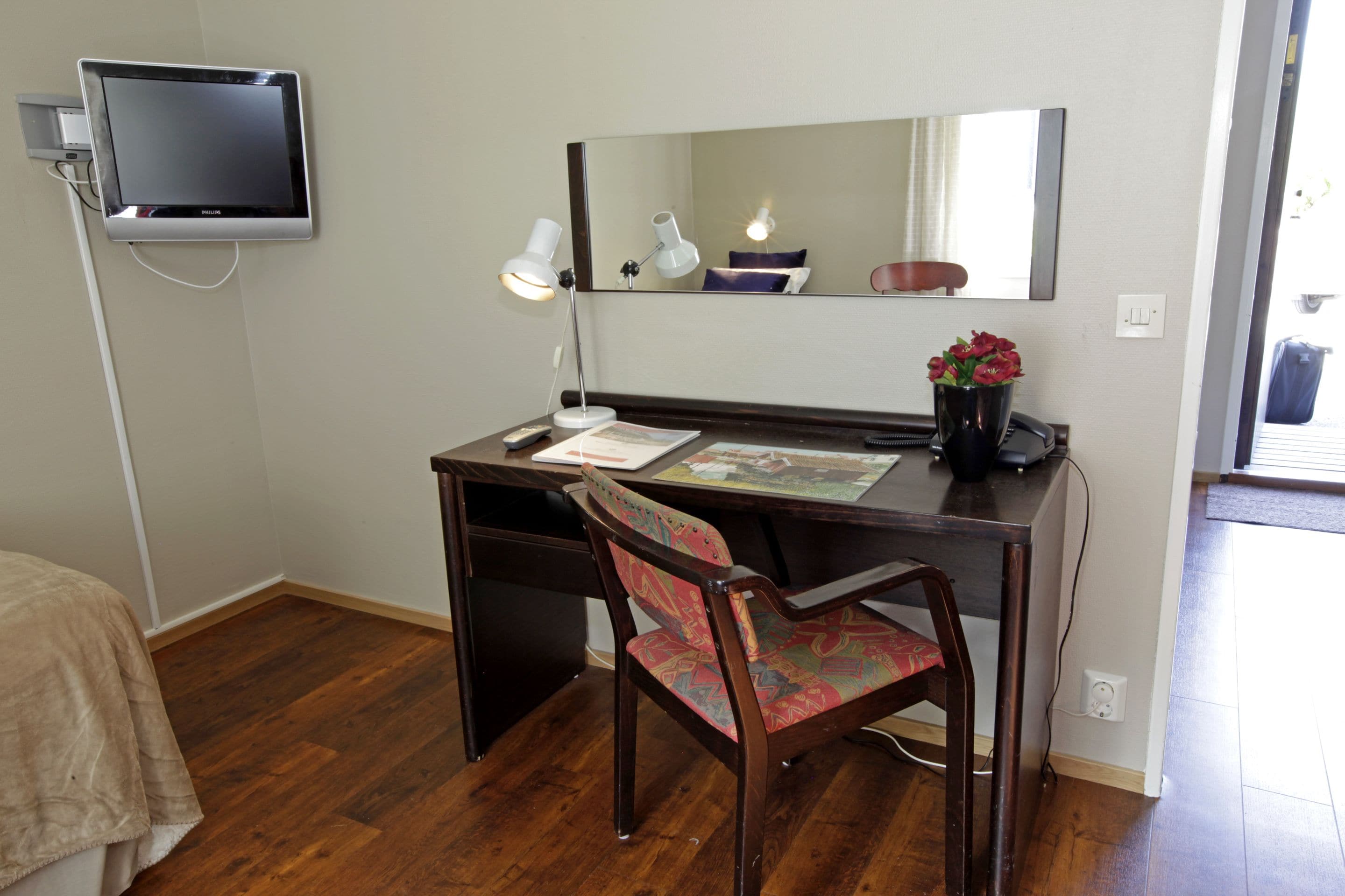 A hotel room with a wooden desk, a chair, a flat-screen TV, a desk lamp, a mirror, and a small potted plant.