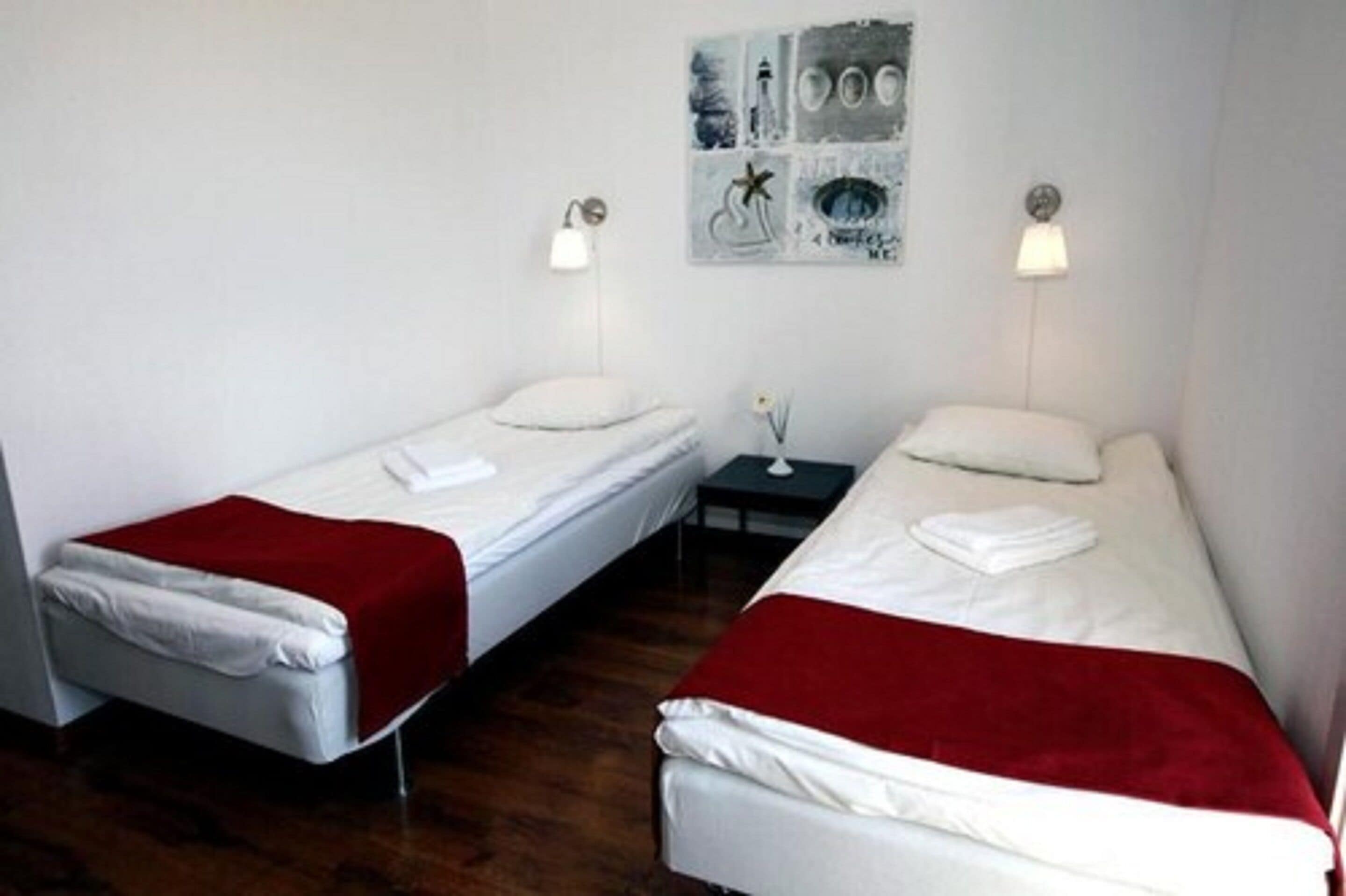 A room with two single beds, each with a white mattress and red blanket, a small table with a lamp, and a wall art piece.