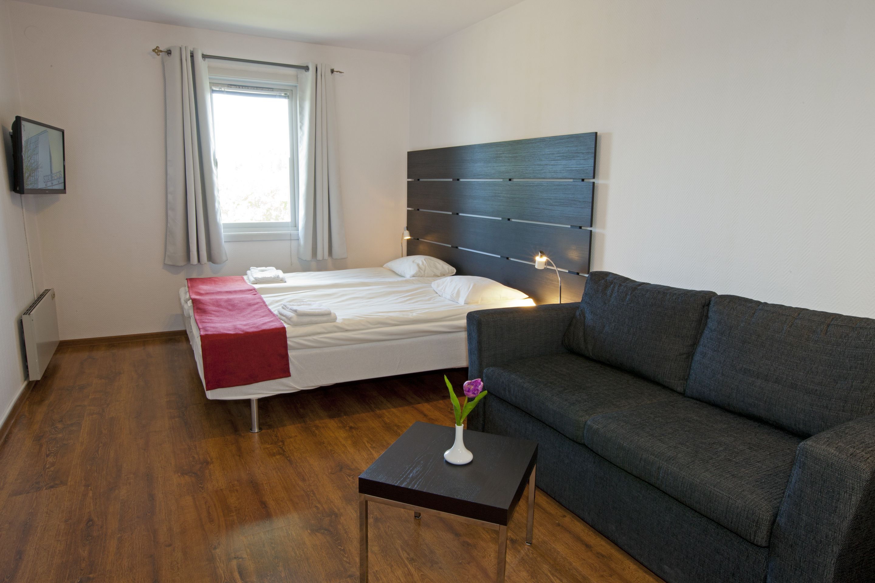 Moderni hotellihuone, jossa on sänky, harmaa sohva, pieni pöytä maljakolla ja ikkuna, jossa on verhot.