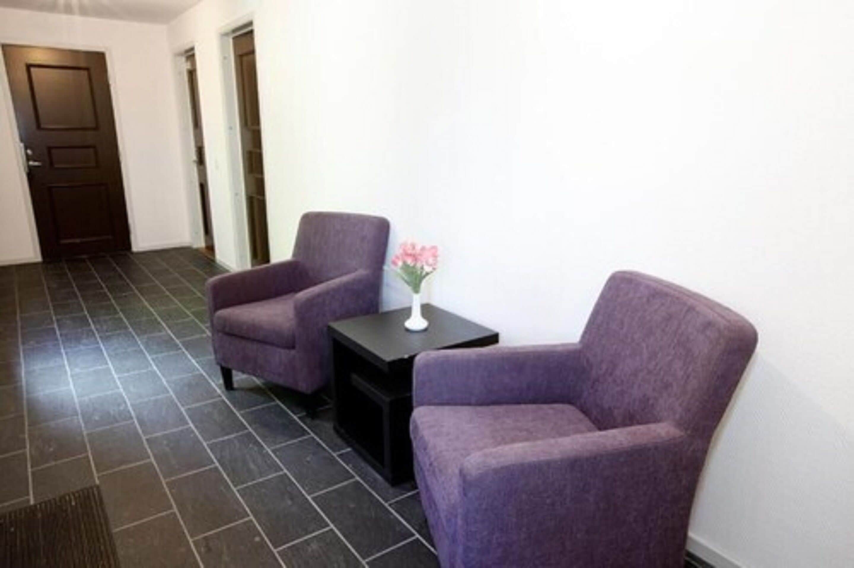 Una habitación con dos sillones morados, una pequeña mesa negra con un florero y un piso de baldosas oscuras.