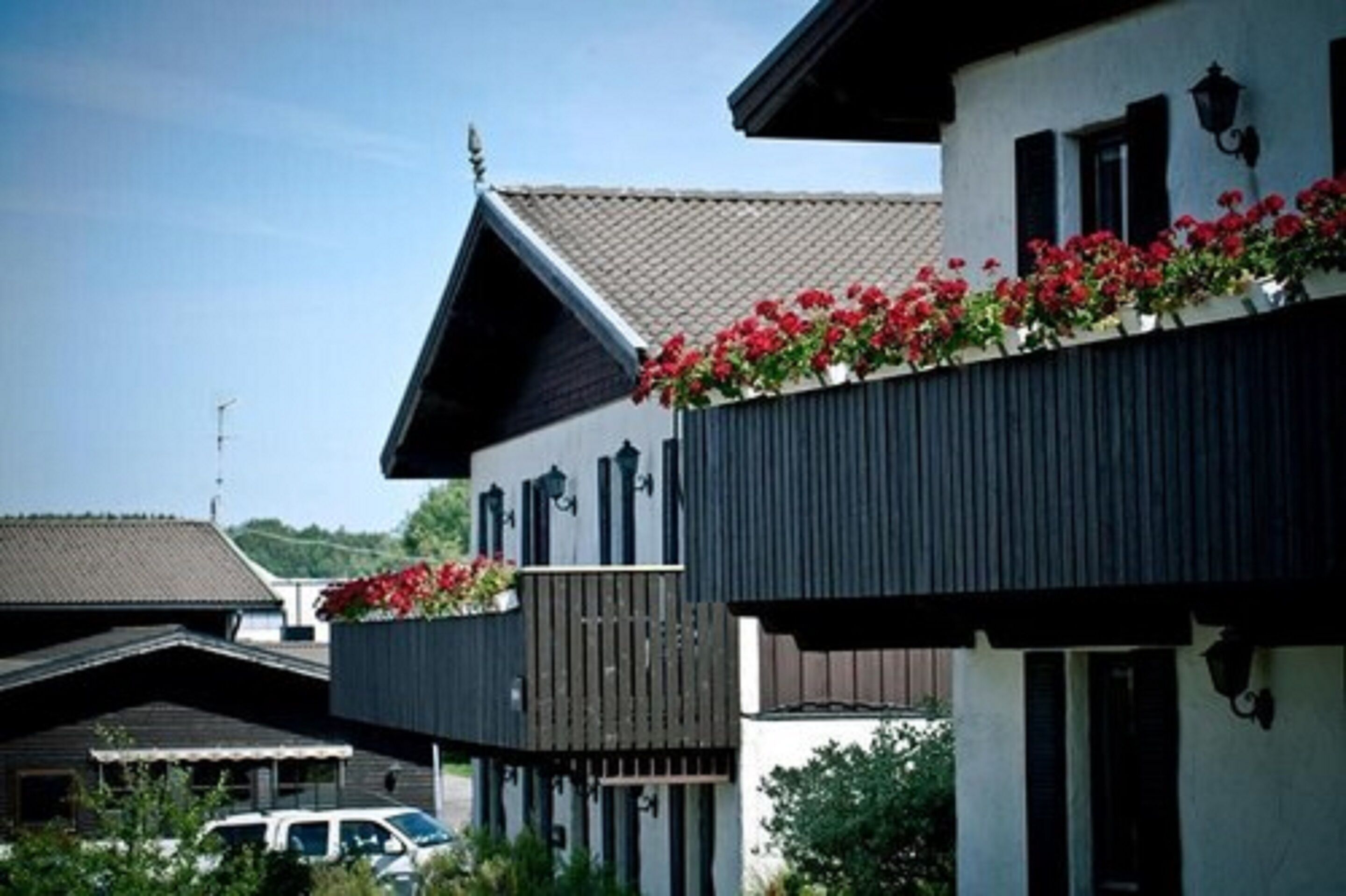 Un edificio de dos pisos con un balcón decorado con flores rojas, un techo inclinado y una antena parabólica visible.