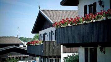 Un edificio de dos pisos con un balcón decorado con flores rojas, un techo inclinado y una antena parabólica visible.