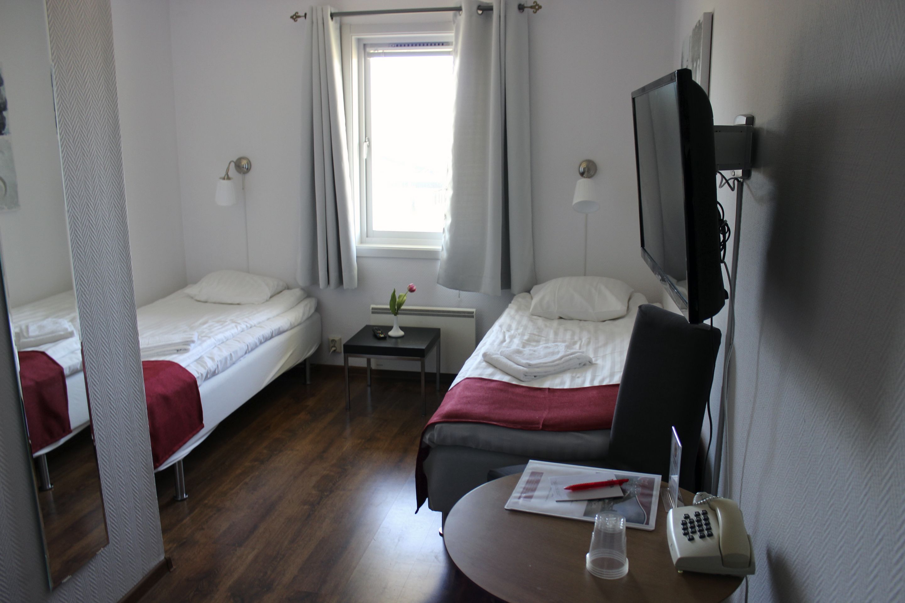 Habitación de hotel con dos camas individuales, una mesita con un teléfono y un vaso, y un televisor fijado a la pared.