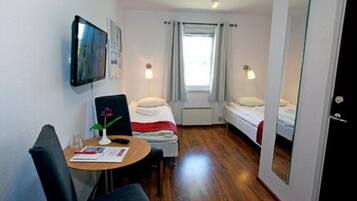 Ein kleines Hotelzimmer mit einem Einzelbett, einem Tisch mit Stuhl, einem an der Wand befestigten Fernseher und einem Fenster mit Vorhängen.