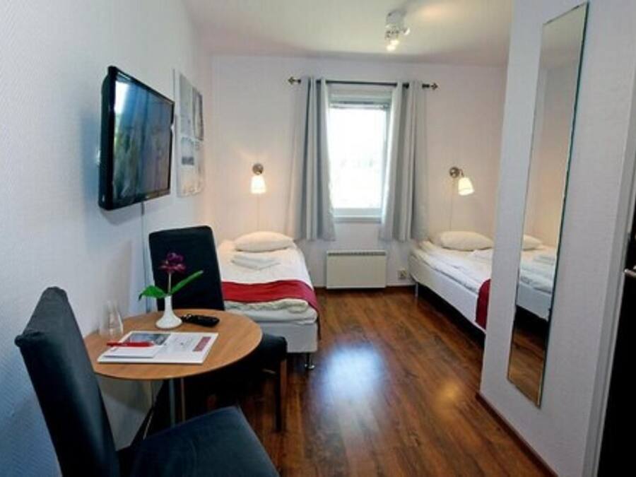 Habitación de hotel compacta con una cama individual, una mesita pequeña con una silla, un televisor montado en la pared y una ventana con cortinas.