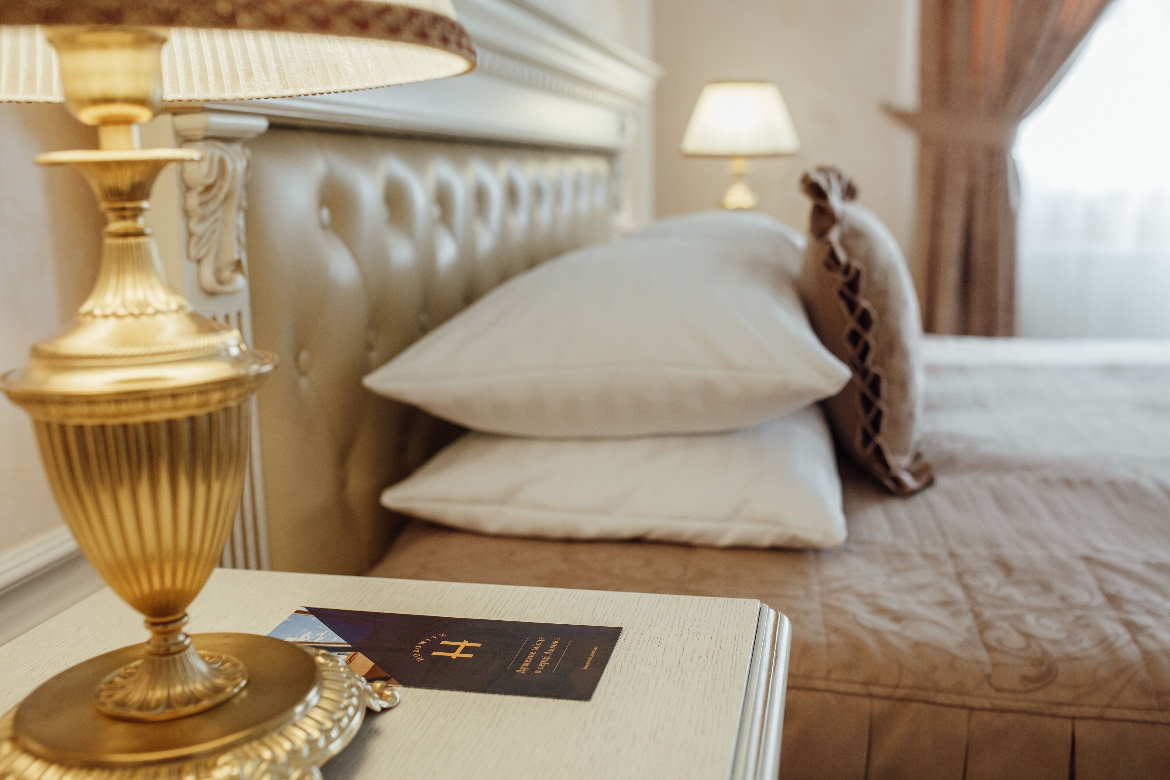 Una habitación de hotel con cama, almohadas, una lámpara y una tarjeta sobre la mesita de noche.