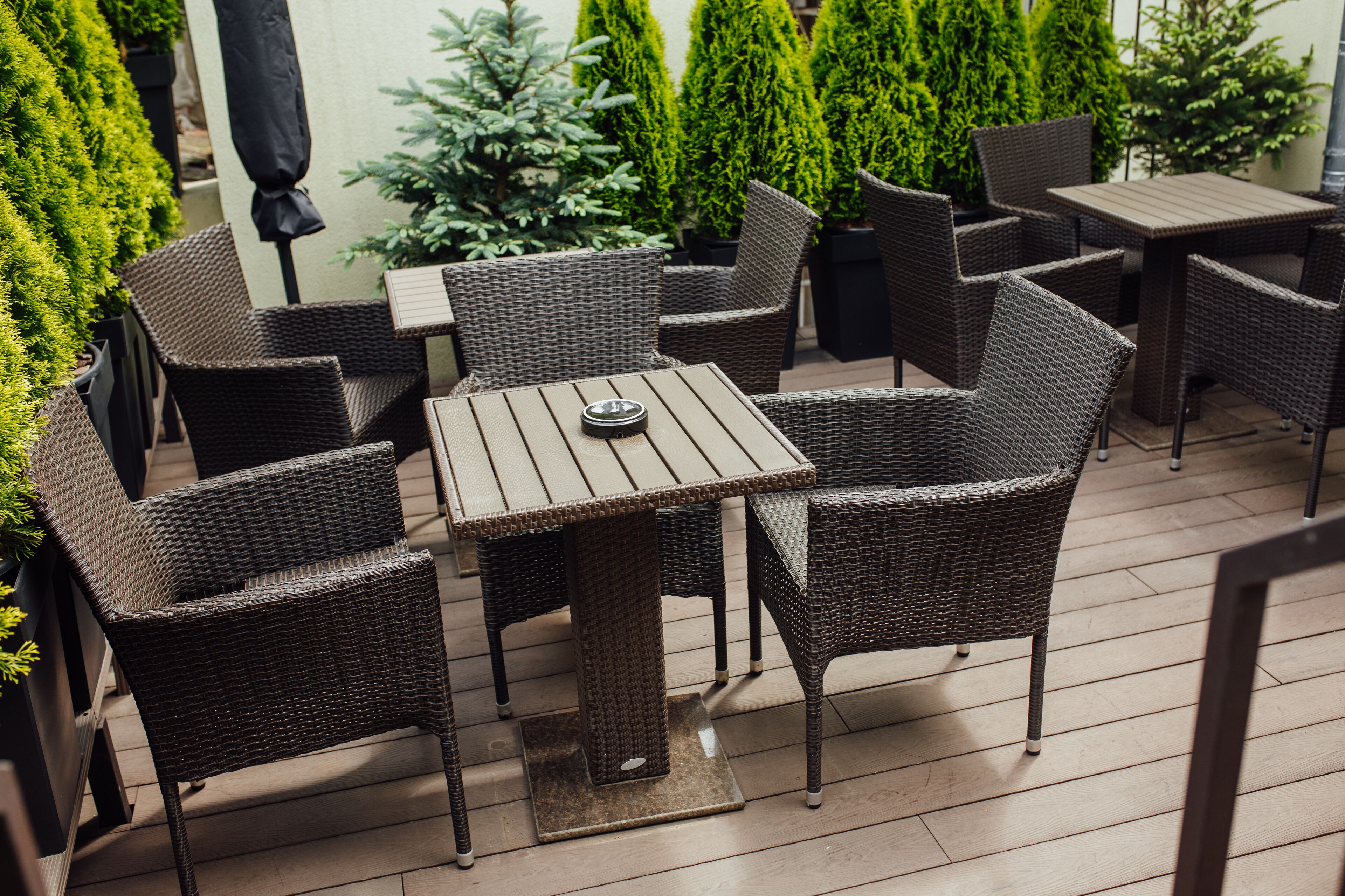 An outdoor patio area with wicker furniture, including a table and chairs, surrounded by potted plants and a wooden deck.