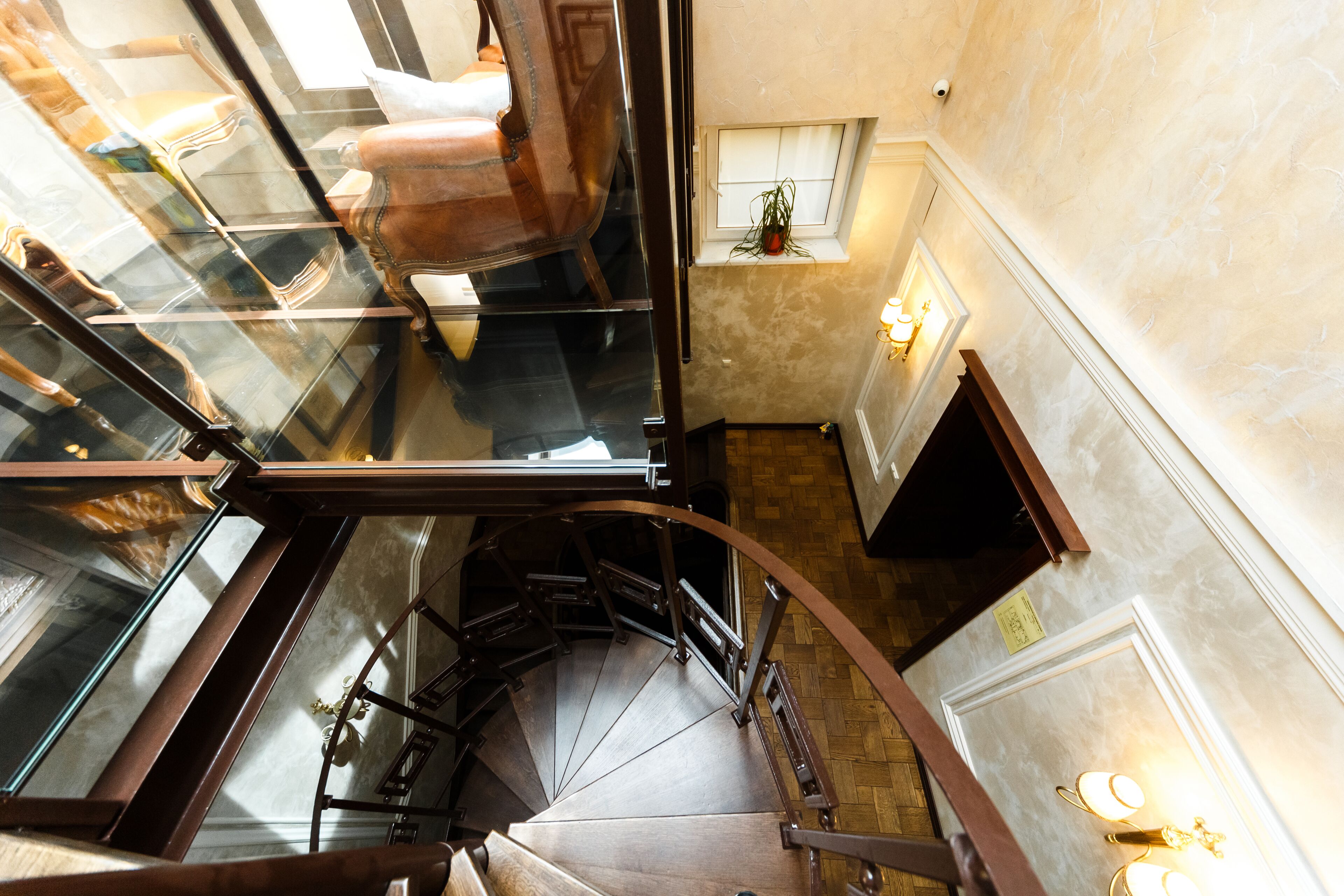 Una escalera de caracol con barandillas de vidrio y vistas a una habitación con suelo de madera y una ventana con planta.