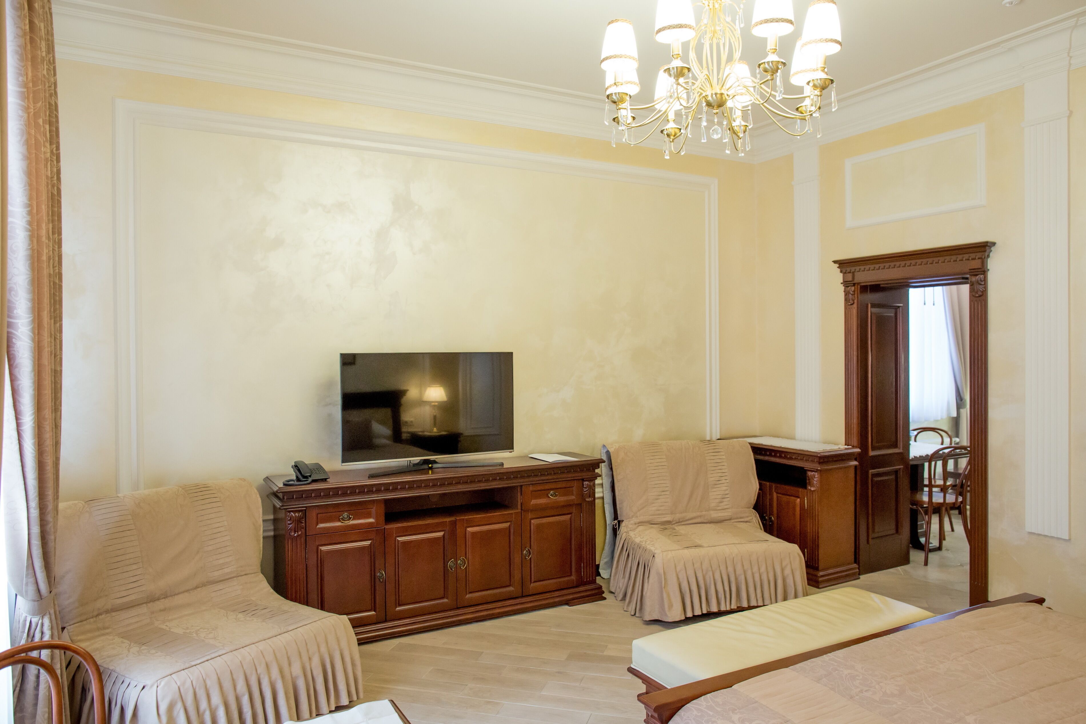 A hotel room with a large bed, two armchairs, a wooden cabinet, a television, and a chandelier.