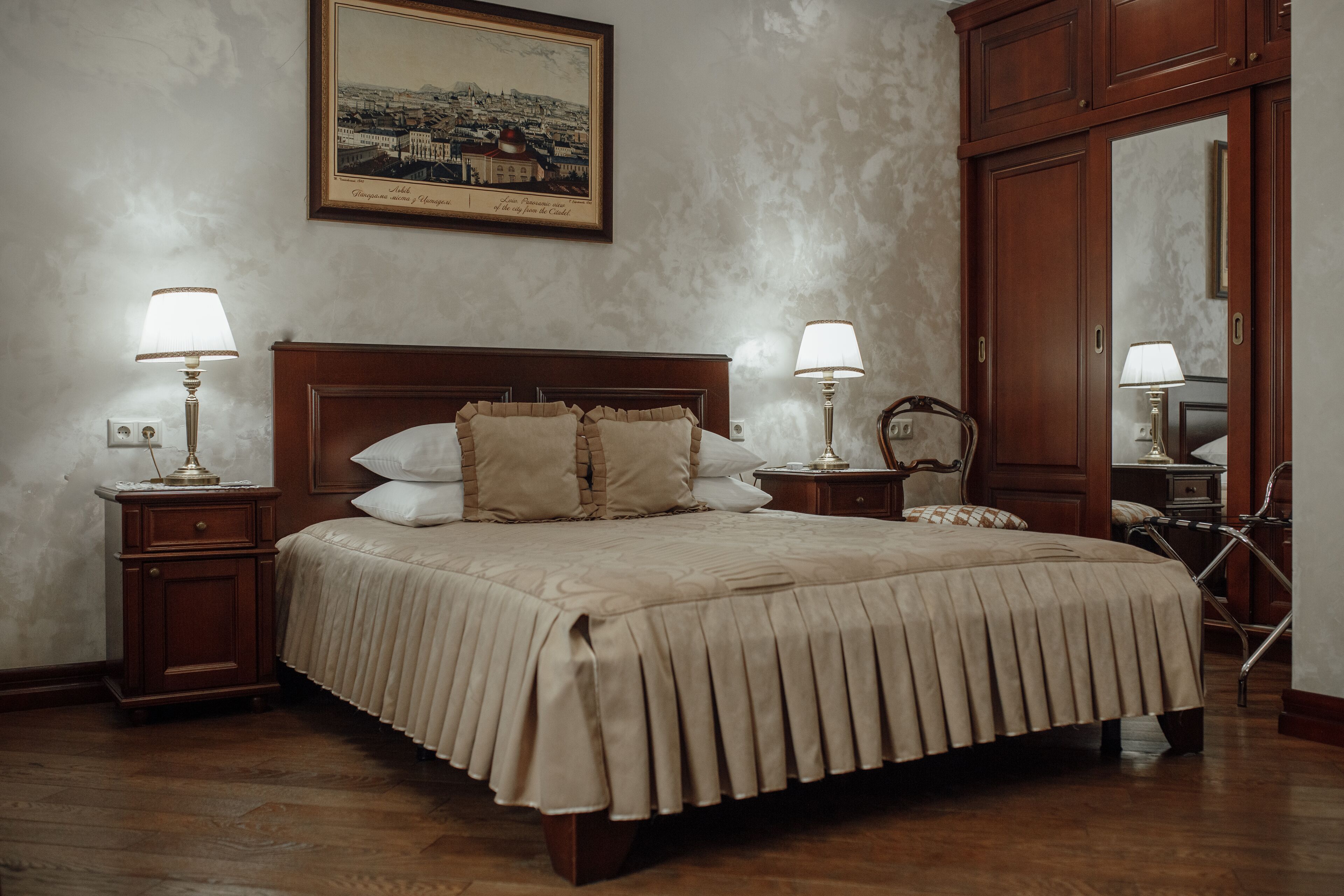Una cama bien hecha con cabecera de madera, dos mesitas de noche con lámparas, un espejo grande y una fotografía enmarcada de un paisaje urbano en la pared.