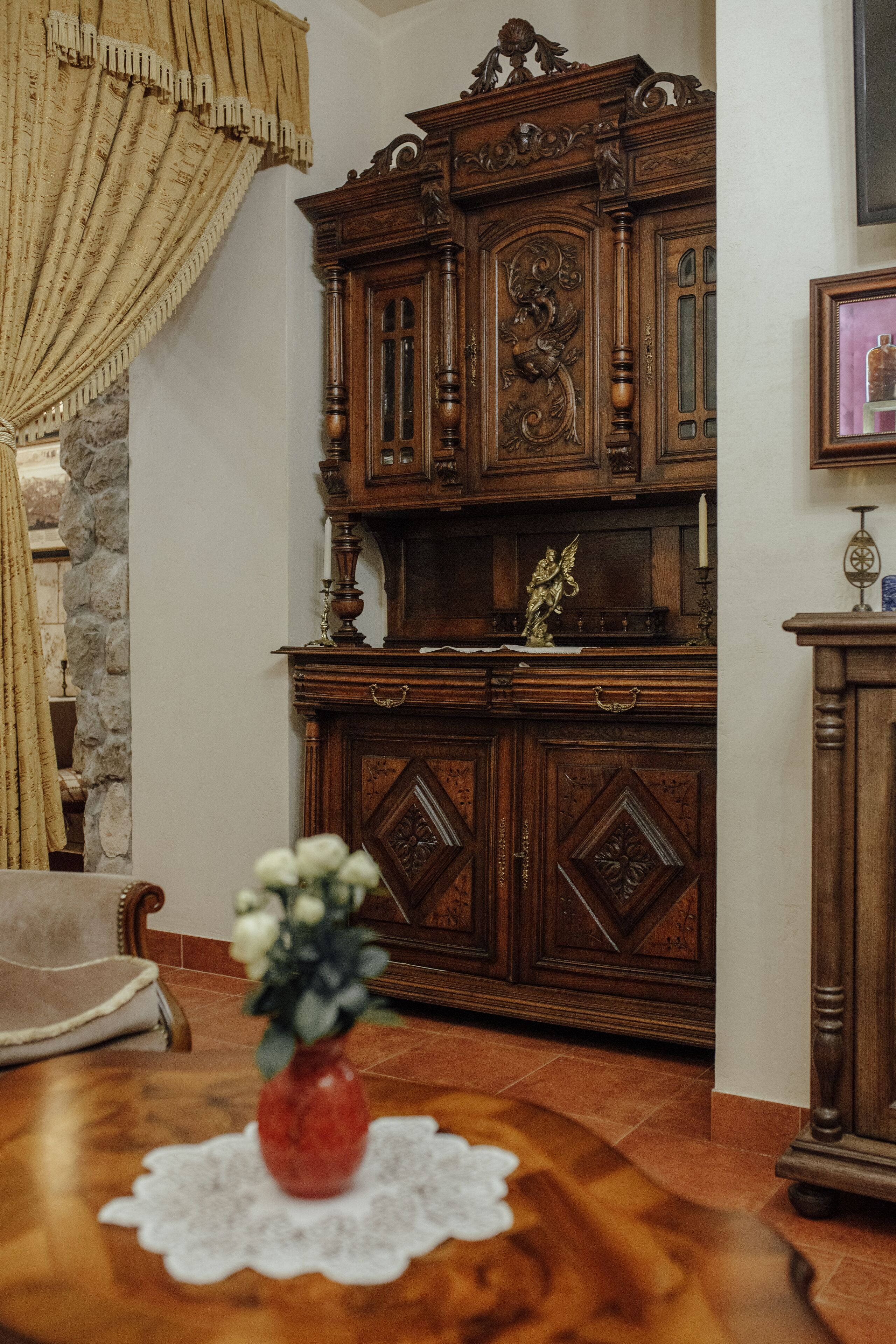 Un aparador de madera con intrincados grabados, un jarrón con flores sobre una mesa, y una pared de piedra.