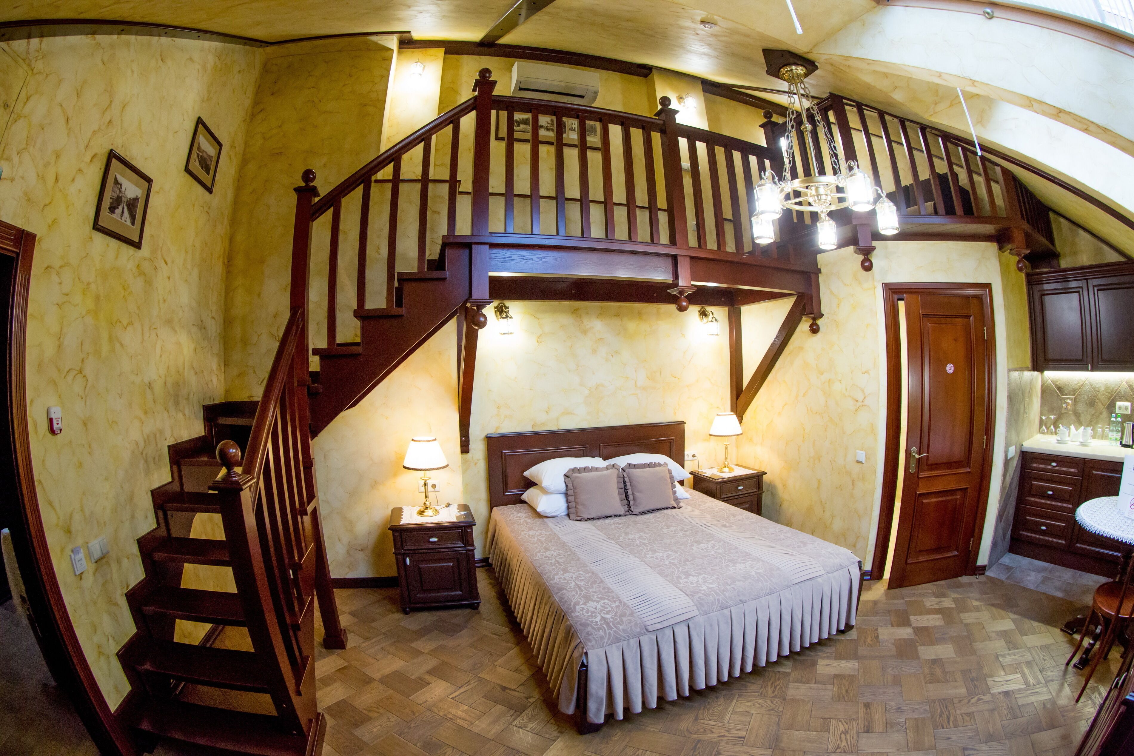 A bedroom with a wooden staircase, a bed with a patterned bedspread, two bedside lamps, and a small table with a chair.